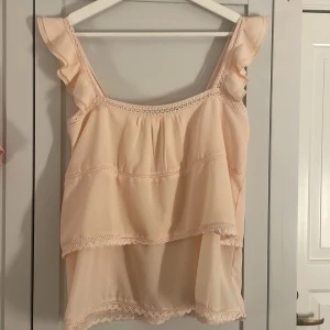 Beige ärmlös topp med volangdetaljer - Säljer en söt beige ärmlös topp med volangdetaljer. Toppen har en luftig passform och är dekorerad med spets längs kanterna. Perfekt för sommardagar eller en avslappnad festkväll. Passar bra till både jeans och kjol!