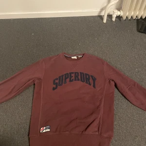 Vinröd tröja från Superdry - Säljer en vinröd tröja från Superdry med tryck på både fram- och baksidan. Säljes pga inte använder så den är i bra skick