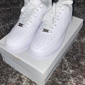 Vita Nike Air Force 1 - Säljer ett par klassiska vita Nike Air Force 1 i nytt skick. De har en stilren design med snörning och perforerade detaljer för extra ventilation. Perfekta för både vardag och fest! Kommer i originalkartong. OBS* Storlek 42,5