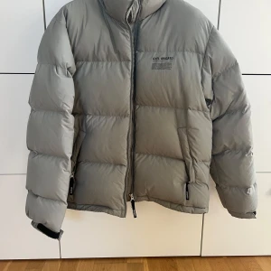 Grå pufferjacka från Axel Arigato - Säljer en snygg grå pufferjacka från Axel Arigato i mycket bra skick. Jackan är perfekt för kalla dagar med sin tjocka vaddering och dragkedja framtill. Den har en stilren design med hög krage och långa ärmar. Passar perfekt för både höst och vinter!