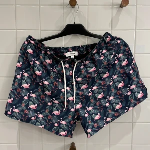 Mönstrade badshorts med flamingos - storlek L - Säljer ett par coola badshorts från Studio Total med ett färgglatt flamingomönster. De är mörkblå med rosa flamingos och gröna blad, perfekt för sommaren! Shortsen har en bekväm passform med snörning i midjan.