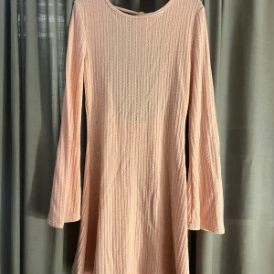 Ljusrosa stickad klänning - Superfin ljusrosa stickad klänning med långärmad design och en snygg öppen rygg med knytning. Perfekt för både vår och höst, och den har en härlig ribbad textur som ger en extra touch. Klänningen är i mycket bra skick och passar perfekt för en mysig dag ute eller en avslappnad fest.