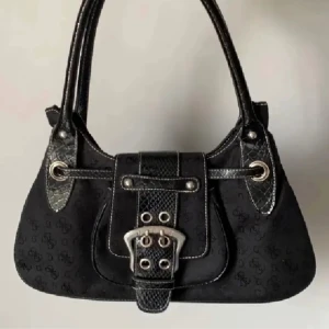 Guess handväska med bälte - Snygg svart vintahe Guess handväska med coola bältesdetaljer och silverfärgade spännen. Väskan har ett diskret mönster och är perfekt för både vardag och fest. Den har en praktisk dragkedja och är i mycket bra skick. Perfekt för att ge din outfit en stilfull touch!