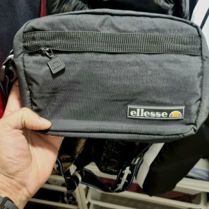 Svart axelväska från Ellesse - Säljer en snygg svart axelväska från Ellesse i mycket bra skick. Väskan har en praktisk dragkedja och en liten ficka framtill för extra förvaring. Perfekt för vardagsbruk eller när du är på språng. Enkel och stilren design som passar till det mesta.