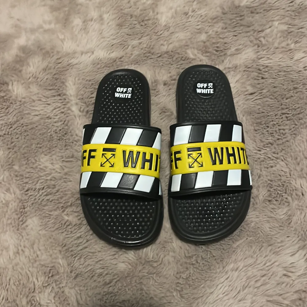 Snygga svarta sandaler från Off-White med coola gula och vita detaljer. De har en bred rem med Off-White-loggan och ett randigt mönster. Perfekta för sommaren och att glida runt i stan med. Sulan är mönstrad för bra grepp. Passar både till stranden och som en casual look.. Övrigt.