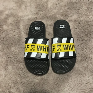 Svarta sandaler från Off-White - Snygga svarta sandaler från Off-White med coola gula och vita detaljer. De har en bred rem med Off-White-loggan och ett randigt mönster. Perfekta för sommaren och att glida runt i stan med. Sulan är mönstrad för bra grepp. Passar både till stranden och som en casual look.