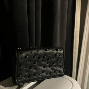 Svart axelväska med nitar - Snygg svart axelväska i skinn med coola nitar över hela framsidan. Väskan har en kedja som axelrem vilket ger den en edgy look. Perfekt för en utekväll eller fest! 🖤