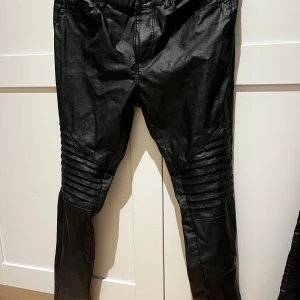Svarta skinnbyxor med detaljer - Snygga svarta skinnbyxor i syntetmaterial med coola ribbade detaljer på knäna. De har en tight passform och är perfekt för en kväll ute eller en rockig look. Byxorna har fickor både fram och bak, vilket är praktiskt. De är i mycket bra skick och redo att bäras till nästa fest!