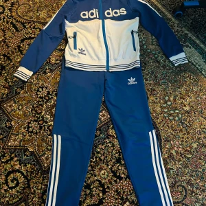 Blå och vit träningsdräkt från Adidas för barn - Säljer en snygg blå och vit träningsdräkt från Adidas i barnstorlek XL. Tröjan har dragkedja och två fickor med dragkedjor. Byxorna har de klassiska tre ränderna längs sidorna. Perfekt för träning eller en avslappnad dag. 🏃‍♂️