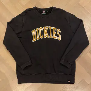 Säljer en svart sweatshirt från Dickies i storlek XL. Tröjan har ett stort gult och vitt tryck med märkesnamnet på framsidan. Den är i bra skick och perfekt för både höst och vinter. Passar till en casual stil och är superbekväm att ha på sig. Perfekt för dig som gillar streetwear!