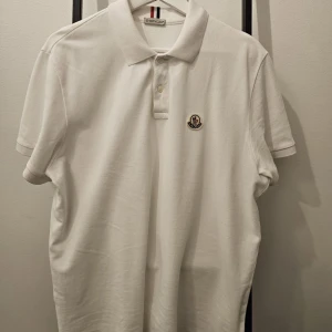 Vit pikétröja från Moncler - Säljer en stilren vit pikétröja från Moncler. Tröjan har en klassisk krage med knappar och Monclers ikoniska logga broderad på bröstet. Perfekt för en avslappnad men ändå snygg look. Passar både till vardags och fest!
