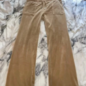 Beige byxor från Juicy Couture - Säljer ett par snygga beige byxor från Juicy Couture i storlek M. De är gjorda i ett mjukt och glansigt material som känns lyxigt. Byxorna har en loose passform och är perfekta för en avslappnad stil. De har fickor både fram och bak med knappar som detaljer. Perfekta för höst och vår! (Pris kan diskuteras)