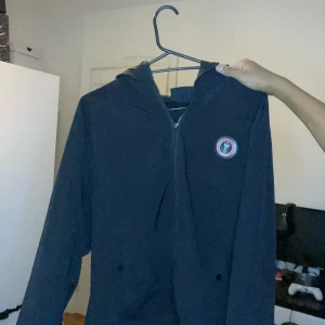 Vindjacka hoodie från Trapstar - Säljer en svart hoodie från Trapstar med dragkedja och huva. Den har en cool logga på bröstet och texten 'Trapstar' i rött på ryggen. Perfekt för höst och vår, och den är i mycket bra skick. Passar både till vardags och chill.