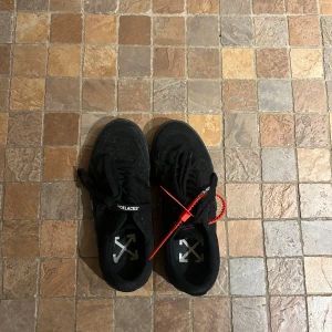Svarta sneakers med röd detalj OFF WHITE - Säljer ett par svarta sneakers med coola detaljer. De har en röd tagg och texten 'SHOELACES' på skosnörena. Perfekta för en stilren look med en twist. Använda men i bra skick. Passar till både vardag och fest!