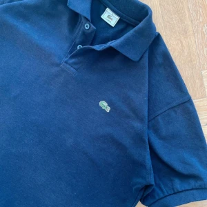 Mörkblå pikétröja från Lacoste - Säljer en klassisk mörkblå pikétröja från Lacoste. Den har en broderad krokodil på bröstet och är i bra skick. Perfekt för både vardag och lite finare tillfällen. Passar bra till jeans eller chinos! Den är i storlek 6 eller L, Öppen för prisförslag!