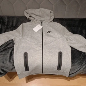 Grå nike tech - Säljer en grå  Nike tech i nytt skick. Den har en dragkedja framtill och två fickor med svarta detaljer. Perfekt för höst och vår, med en bekväm passform och huva för extra värme. En stilren och praktisk tröja för vardagsbruk.