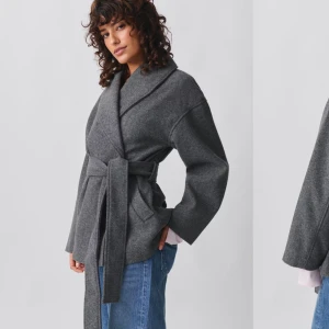 Grå kappa med knytband - Säljer en stilren grå kappa i ull med knytband i midjan. Perfekt för höst och vår! Den har en loose passform och långa ärmar, vilket gör den både bekväm och trendig. Kappan är i mycket bra skick och passar perfekt för både vardag och fest.