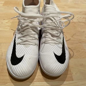 Vita futsalskor från Nike - Säljer ett par vita Nike futsalskor i mycket bra skick. Skorna har en snygg svart Nike-logga och röda detaljer på sidan. De är designade för inomhusbruk med en slitstark sula och har snörning för perfekt passform. Perfekta för fotbollsträning eller match! Använda ändast 4-5 gånger