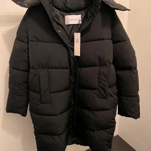  Calvin Klein vinterjacka  - Säljer en snygg och varm svart dunjacka från Calvin Klein. Jackan är i nytt skick med etikett kvar. Den har en pufferstil med långa ärmar och en praktisk huva. Perfekt för kalla vinterdagar! Calvin Klein MODERN PADDED COAT Size L 