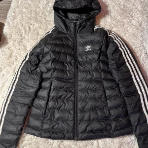 Svart dunjacka från Adidas - Säljer en snygg svart dunjacka från Adidas i storlek XS. Jackan har en huva och klassiska vita ränder längs ärmarna. Den är perfekt för kyliga dagar och har en dragkedja framtill. Adidas-loggan syns tydligt på bröstet. Perfekt för både vardag och sportiga aktiviteter!