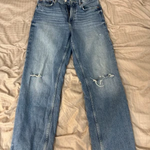 Blå jeans med slitningar från zara - Säljer ett par snygga blå jeans med slitningar på knäna. De har en klassisk femficksdesign och en bekväm passform. Perfekta för en avslappnad vardagslook. Passar bra till både sneakers och kängor!