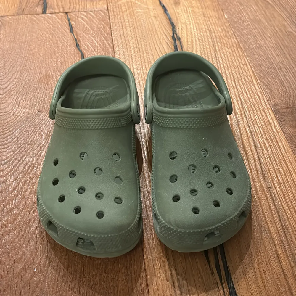 Säljer ett par olivgröna Crocs i syntetmaterial. Perfekta för sommarens alla äventyr med sin bekväma passform och ventilerande hål. De har en justerbar hälrem för extra stöd. Använd dem på stranden eller i trädgården för maximal komfort.. Skor.