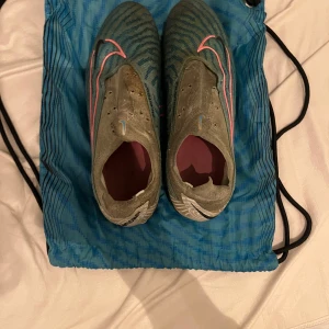 Gröna fotbollsskor från Nike - Säljer ett par blå Nike fotbollsskor med rosa detaljer. Skorna har en cool mönstrad yta och är perfekta för fotbollsträning. De är i dåligt skick så billigt pris, och kommer med en praktisk blå förvaringspåse. Original pris 3300