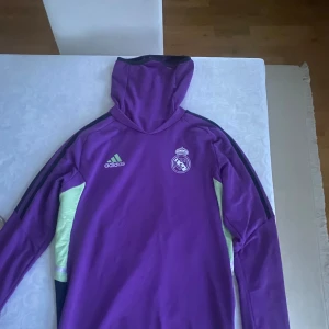 Lila hoodie från Adidas med Real Madrid-logga - Säljer en snygg lila hoodie från Adidas med Real Madrid-logga på bröstet. Den har en cool design med svarta och ljusgröna detaljer. Perfekt för fotbollsfans som vill ha något bekvämt och stiligt. Passar bra till både träning och vardag. Storlek xs, passar 165-175. Nypris 1599kr