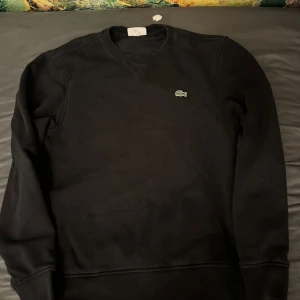Svart tröja från Lacoste - Säljer en klassisk svart tröja från Lacoste i storlek M. Den har en stilren design med det ikoniska krokodilmärket på bröstet. Perfekt för både vardag och lite finare tillfällen. Passar bra till jeans eller chinos!