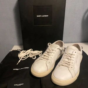 Vita sneakers från Saint Laurent - Säljer ett par stilrena vita sneakers från Saint Laurent i skinn. De har en klassisk design med snörning och en bekväm sula. Perfekta för både vardag och festliga tillfällen. Skorna är i använt skick och kommer med originalförpackning,dustbags och extra skosnören.