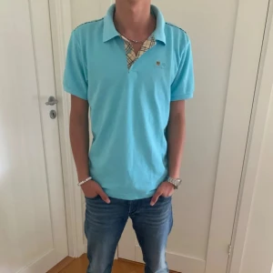 Ljusblå piké från Burberry - Säljer en snygg ljusblå piké från Burberry i mycket bra skick. Tröjan har korta ärmar och en klassisk krage med det ikoniska Burberry-mönstret på insidan. Perfekt för sommardagar eller en avslappnad vårlook. Passar både till jeans och chinos! Skick 9,5/10 inga skador eller defekter , nypris  1495