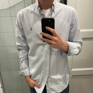 Randig skjorta från Ralph Lauren - Säljer en stilren och klassisk skjorta från Ralph Lauren. Den är vit med ljusblå ränder och har en broderad logga på bröstet. Den är endast använd 1 gång! Skjortan är långärmad och har en normal passform, perfekt för både vardag och fest💙 Knapparna framtill ger en snygg och elegant look. Passar bra till jeans eller chinos!