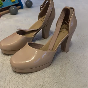 Beige pumps med klack - Snygga beige pumps från Vox i syntetmaterial. De har en hög klack och en platåsula som ger extra höjd och stil. Perfekta för festliga tillfällen eller en utekväll. Skorna har en elegant design med en rem runt ankeln för bättre passform.