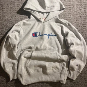Grå hoodie från Champion - Säljer en klassisk grå hoodie från Champion i mycket bra skick. Den har en stor broderad logga på bröstet och en mindre på ärmen. Perfekt för höst och vinter med sin mysiga passform och känguruficka framtill. Huvan har dragsko för justering. Perfekt för chill dagar eller träning! Pris kan diskuteras.
