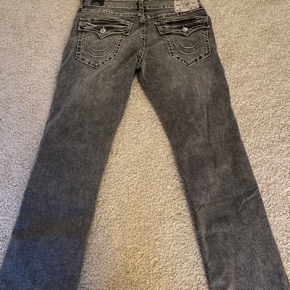 HELT NYA Säljer ett par grå jeans från True Religion i modellen Ricky Big T Flap. De har en straight fit och är i en snygg grå tvätt med slitna detaljer. Bakfickorna har coola sömmar och knappar. Perfekta för en avslappnad stil!. Farkut & Housut.