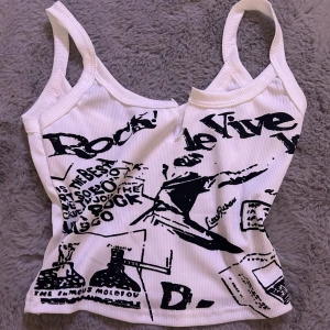 Vit ribbad linnetopp med tryck - Säljer en cool vit ribbad linnetopp med svart tryck. Toppen har en djup urringning och breda axelband. Perfekt för en avslappnad och trendig look. Passar bra till jeans eller kjol för en casual stil.