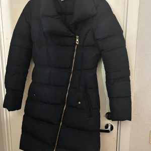 Svart dunjacka från Versace Collection - Säljer en elegant svart dunjacka från Versace Collection. Jackan har en asymmetrisk dragkedja och är långärmad, perfekt för kalla vinterdagar. Den är i mycket bra skick och har en stilren design som passar både till vardags och fest. Storlek är inte specificerad, men den ser ut att ha en normal passform. Nypris 6000kr, Storlek S