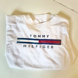 Vit t-shirt från Tommy Hilfiger - Säljer en klassisk vit t-shirt från Tommy Hilfiger med deras ikoniska logga i blått, vitt och rött på bröstet. Perfekt för en avslappnad stil och passar till allt. T-shirten är i bra skick och redo för nya äventyr!