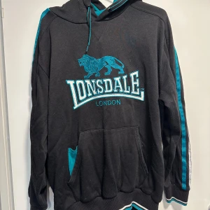 Svart hoodie från Lonsdale - Säljer en svart hoodie från Lonsdale med turkosa detaljer och ett stort tryck på framsidan. Superbekväm och i bra men använt skick! lite nopprig osv därav priset. I storlek XXL men skulle säga att den passar M (unisex) 