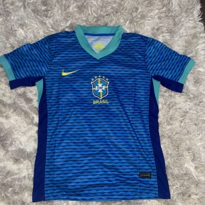 Blå och svart randig Brasilien fotbollströja - Säljer en snygg Brasilien fotbollströja från Nike. Den är blå med svarta ränder och har korta ärmar. Tröjan har en V-ringad krage i ljusblått och det klassiska Brasilien-emblemet på bröstet. Perfekt för fotbollsfans som vill visa sitt stöd för laget!