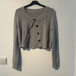 Grå stickad kofta med knappar - Säljer en superfin grå stickad kofta med snygga flätmönster på både fram- och baksidan. Den har långa ärmar och stora, mörka knappar som ger ett fint kontrast. Perfekt för höst och vinter när du vill hålla dig varm och stilren. Koftan är croppad och har ribbade kanter som ger en skön passform. knapptanvänd, lite nopprig men går att få bort💕