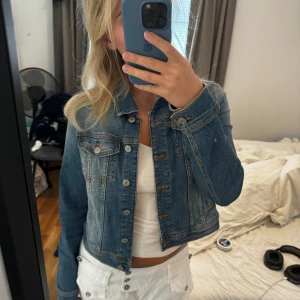 Blå croppad jeansjacka - Säljer en snygg blå jeansjacka med knappar framtill. Jackan har en klassisk jeanslook med två bröstfickor och långa ärmar. Perfekt för en casual stil och passar bra till både vår och sommar.
