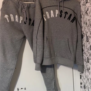 Grå hoodie från Trapstar - Säljer en grå hoodie från Trapstar med stor logga i vitt och svart på bröstet. Den är superbekväm och perfekt för chill dagar. Hoodien har en klassisk känguruficka och justerbar huva. Perfekt för höst och vinter! byxorna ingår 