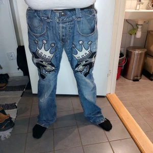 Jeans med coolt tryck - Säljer ett par unika jeans med ett stort tryck av kronor och ansikten på framsidan. De är blå och har en klassisk femficksdesign. Perfekta för dig som vill sticka ut lite extra! Passar bra till vardags eller en avslappnad fest.