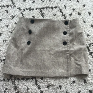 Grå kortkjol med knappar - Säljer en snygg grå kortkjol med svarta knappar framtill. ull kjol. Den har en omlott-design, går att knäppa upp de tre knapparna. 