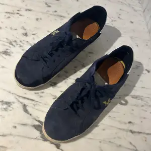 Säljer ett par mörkblå sneakers från Lyle & Scott. Skorna är i mocka och har en guldfärgad logga på sidan. De har snörning och en vit sula som är lite smutsig. Perfekta för vardagsbruk och ger en snygg och avslappnad look.