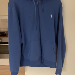 Blå hoodie från Ralph Lauren - Hej, jag säljer en snygg Ralph lauren zip hoodie som är i bra skick och inga defekter, hoodien passar bra med ett par blåa jeans och en vit t shirt enligt mig. Perfekt för höst och sommaren! om ni har några frågor är det bara att fråga