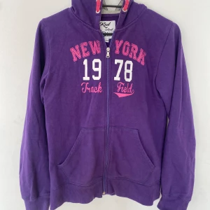 Lila hoodie från Red Spirit - Säljer en snygg lila hoodie från Red Spirit med texten 'New York 1978 Track Field' i rosa och vitt på framsidan. Den har en dragkedja framtill och en mysig huva. Perfekt för höst och vinter! 💜