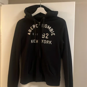 Hoodie från Abercrombie & Fitch - Säljer en mörkblå hoodie från Abercrombie & Fitch med texten 'Abercrombie 92 New York' i vitt på framsidan. Tröjan har dragkedja och en mysig huva med snören. I jättefint skick!!