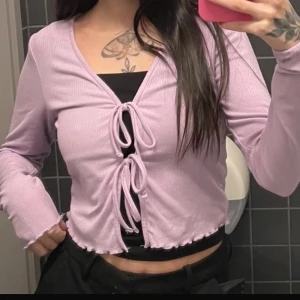 Ljuslila ribbad topp med knyt - Säljer en söt ljuslila ribbad topp med knyt framtill. Toppen har långa ärmar och en v-ringad design. Perfekt för en trendig look!